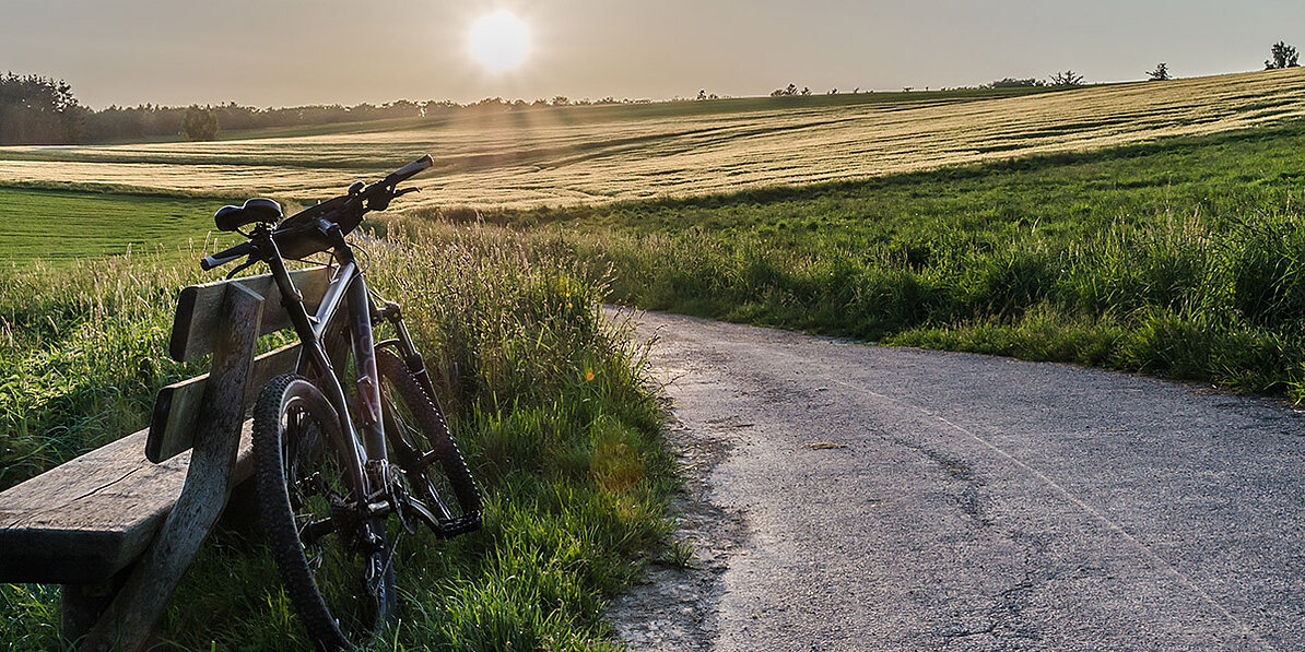 Das Bild zeigt ein Mountainbike, das an einer Holzbank am Rande eines Feldwegs steht, während die Sonne im Hintergrund über einem grünen Feld untergeht.