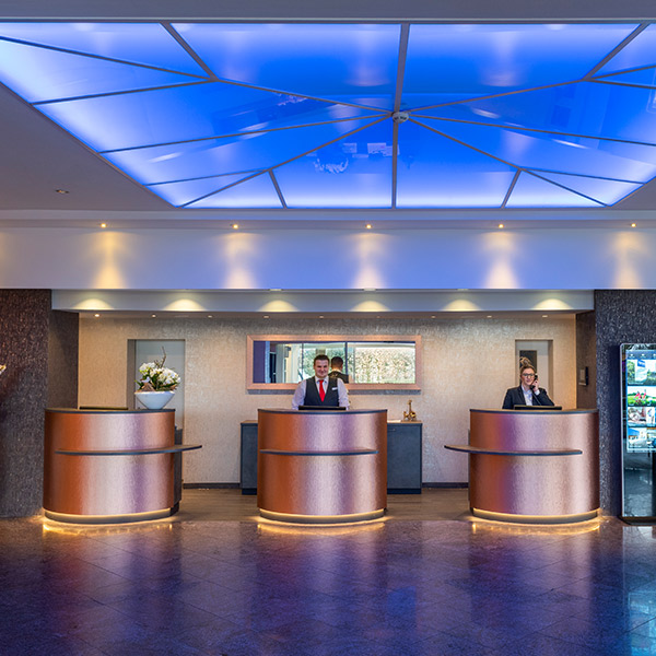 Das Bild zeigt die Rezeption eines modernen Hotels mit zwei Angestellten an den Empfangstresen und einem auffälligen, blau beleuchteten Glasdach.