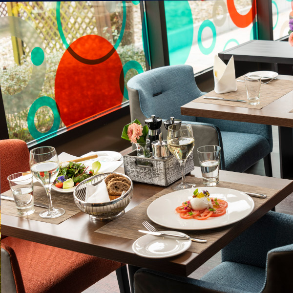 Ein Tisch in einem Restaurant mit Blick auf ein abstraktes Fenster ist mit einem Teller mit Burrata-Salat, Brot und Getränken gedeckt.